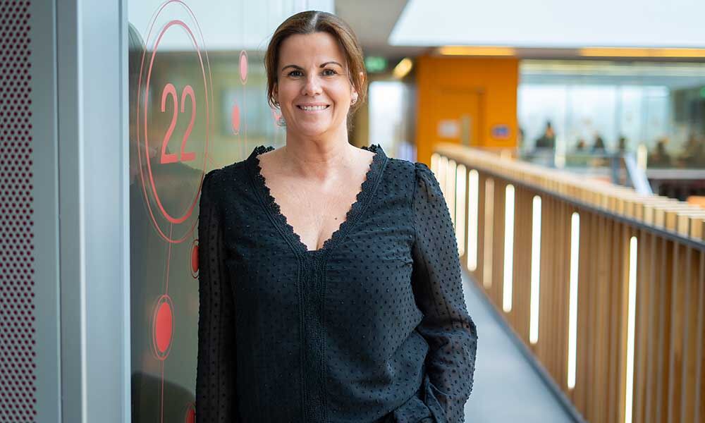 Dark haired woman wearing black top, leaning against glass wall and smiling.